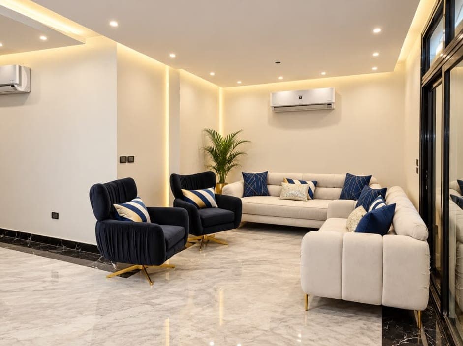 Apartment living room with cream corner sofas, two navy armchairs, a potted palm and polished marble flooring
